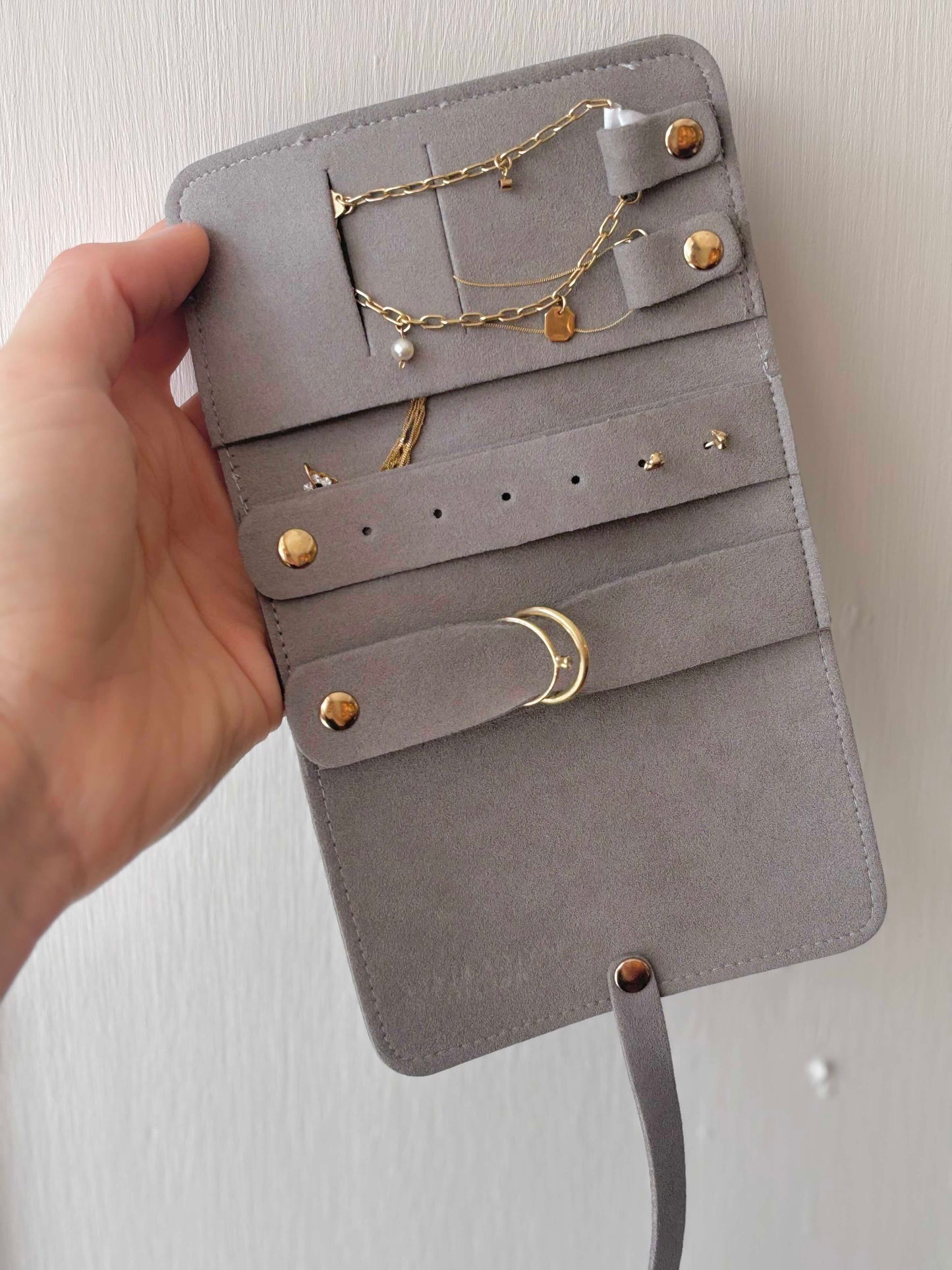 A gray suede jewelry organizer roll-up case held open by a hand, showing gold-tone rings, earrings, and chains stored in its slots against a light wall.