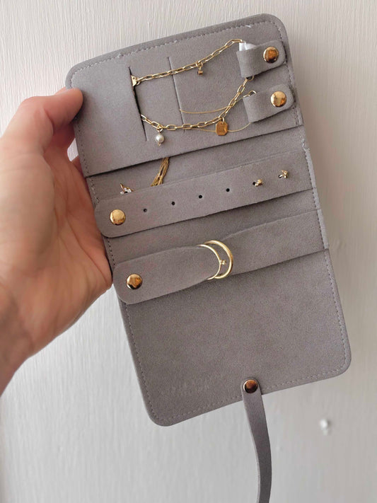 A gray suede jewelry organizer roll-up case held open by a hand, showing gold-tone rings, earrings, and chains stored in its slots against a light wall.