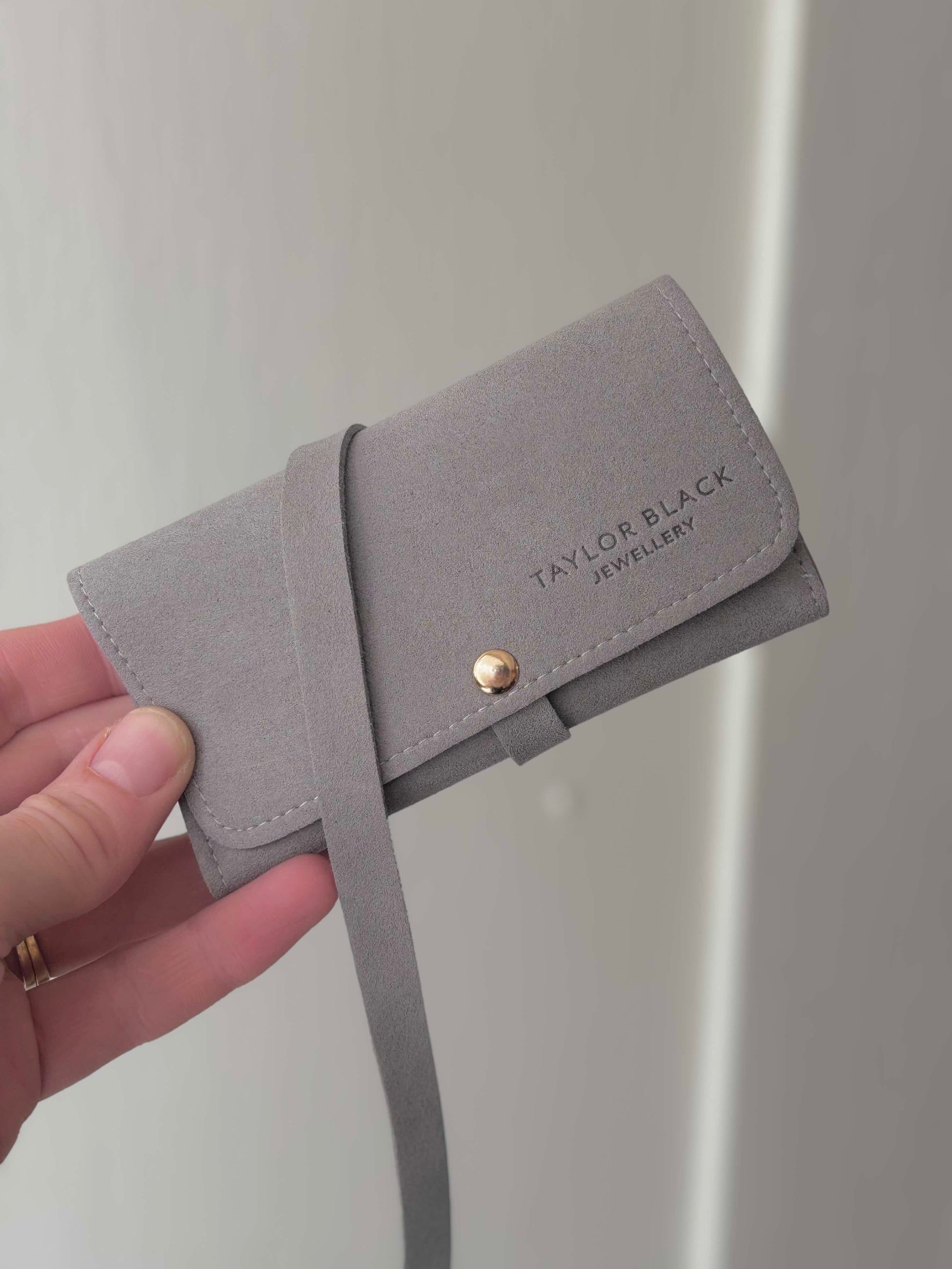 A hand holding a light gray faux-leather jewelry wallet with a strap and a gold snap button. The wallet has 'TAYLOR BLACK JEWELLERY' printed on the front flap.