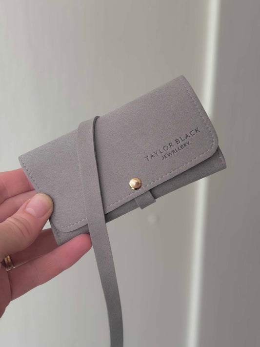 A hand holding a light gray faux-leather jewelry wallet with a strap and a gold snap button. The wallet has 'TAYLOR BLACK JEWELLERY' printed on the front flap.